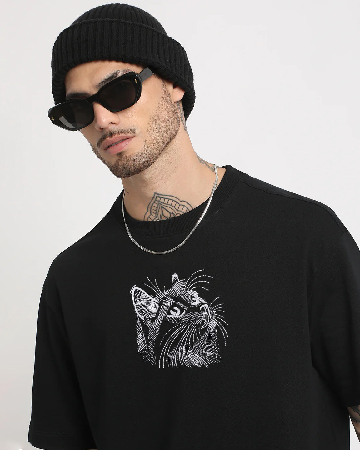 Man wearing a black sweater with a cat graphic and a black beanie and sunglasses on a white background