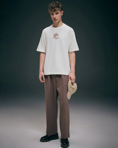 Man wearing a white t-shirt with a logo and brown pants, holding a beige cap against a dark background